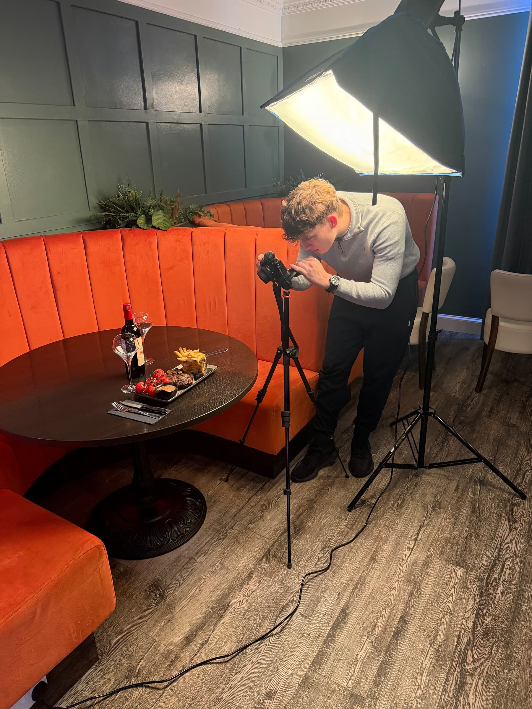 Sean from Jigsaw Studio bent over a camera on a tripod, lining up a steak dinner under a softbox in a restaurant booth